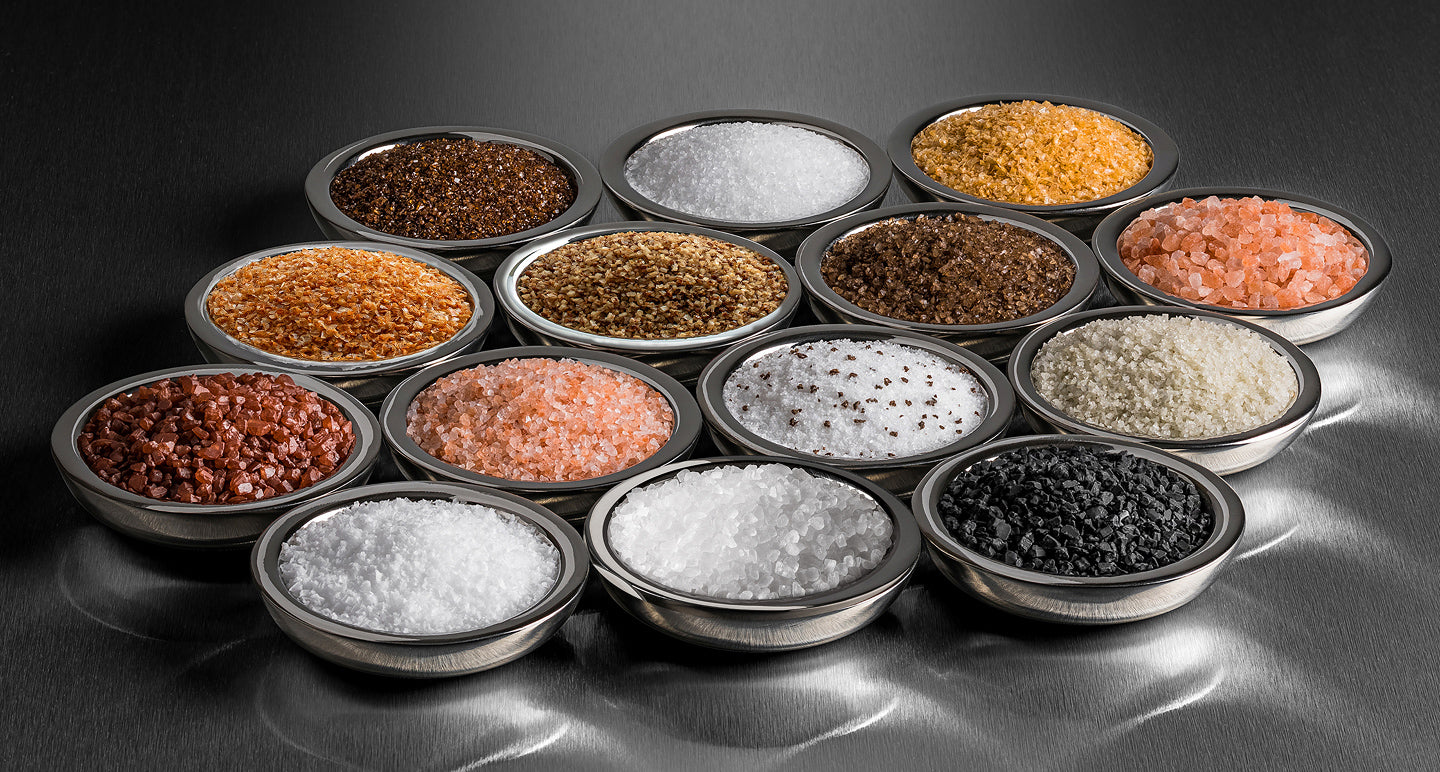 Assorted types of salt in small bowls on a reflective surface with a dark background