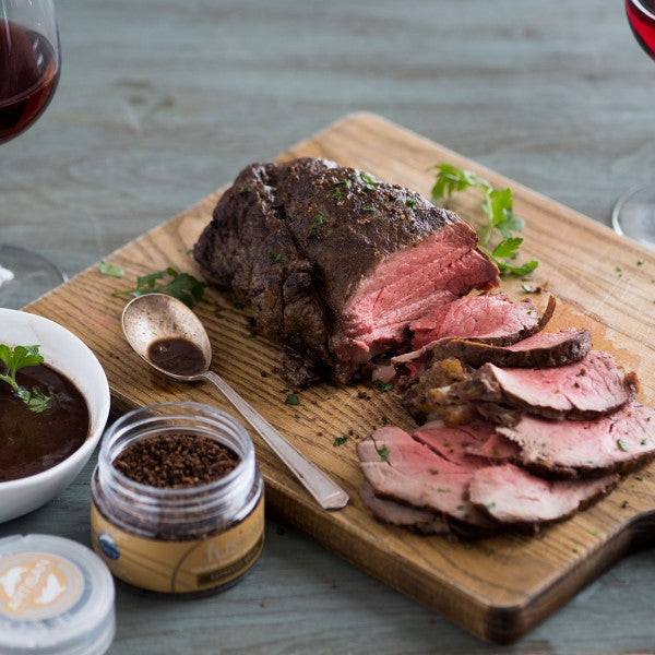 Sliced roast beef on a wooden board with a jar of seasoning, spoon, and bowl of sauce.