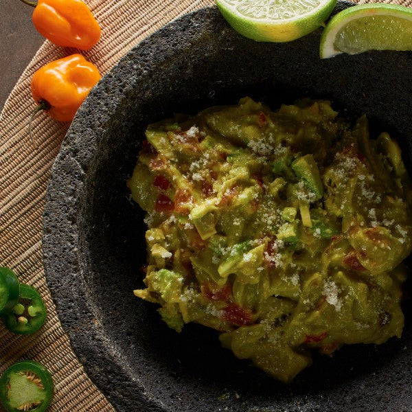 Guacamole in a stone bowl with limes and peppers on a textured surface