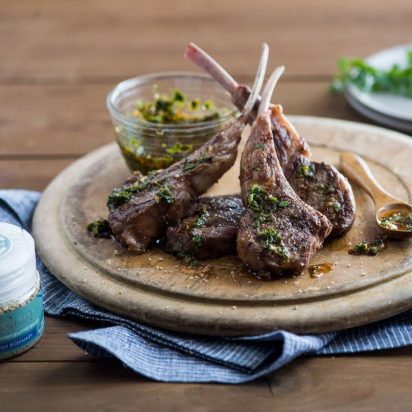 Lamb chops on a wooden plate with a small dish of green sauce on a wooden table.