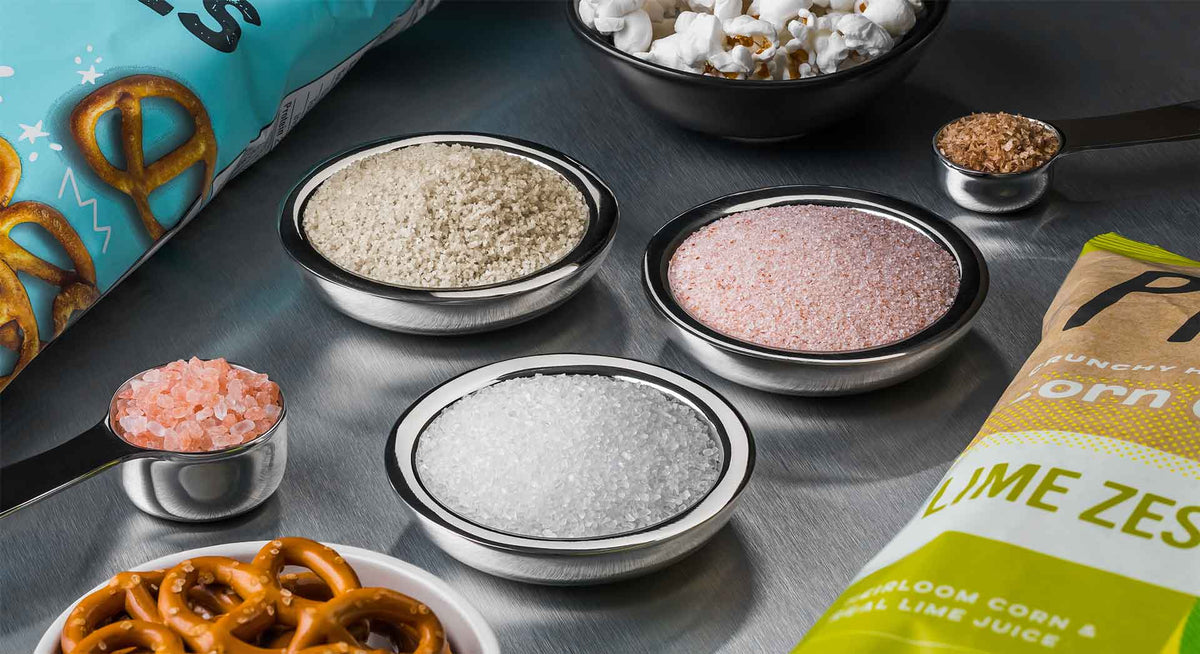Assorted salts in stainless steel bowls next to snacks on stainless steel surface