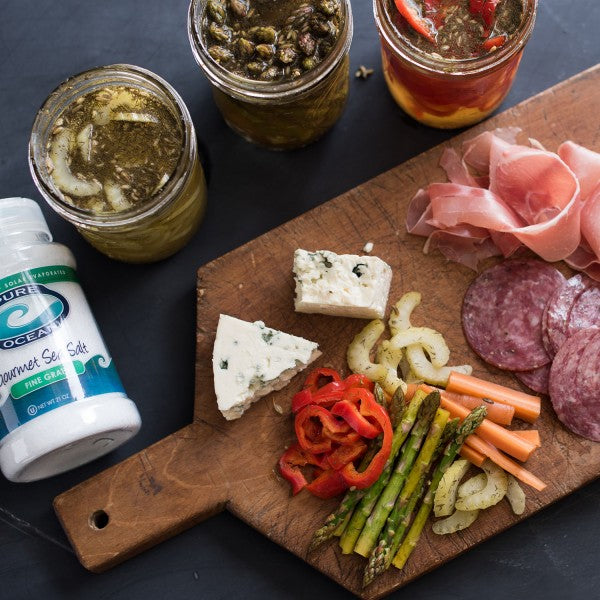 Wooden cutting board with cured meats, cheeses, and vegetables next to jars of pickles and a container of Gourmet Sea Salt.