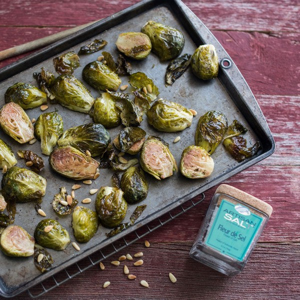 Roasted Brussels sprouts on a baking tray with a container of fleur de sel on a wooden surface.