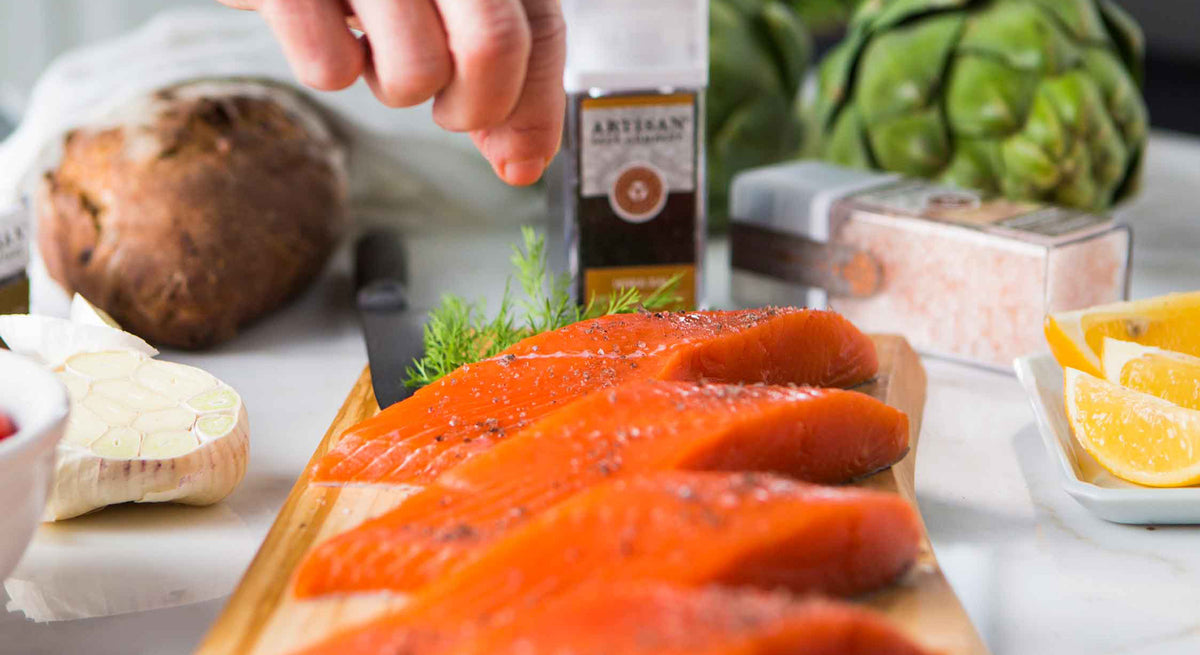 Salmon fillets on a wooden board with a hand holding an Artisan grinder jar