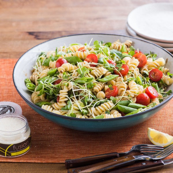 Pasta salad with vegetables in a blue bowl on a wooden table, with a jar of dressing and cutlery beside it.