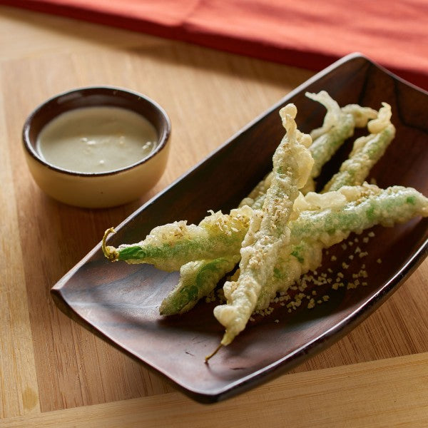 Tempura green beans on a dark plate with a small bowl of dipping sauce on a wooden surface.